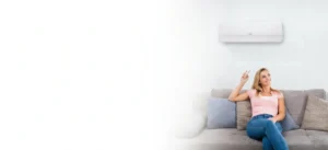 Woman sitting on a gray sofa in a bright living room, holding a remote toward a wall-mounted air conditioner.