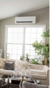 White wall-mounted air conditioner sits above a large window in a bright neutral living room with a tufted beige sofa and green houseplants nearby.