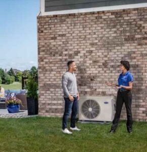 Two men conversing outside a brick building next to an outdoor air conditioner unit.