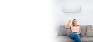 Woman on a gray sofa in a bright living room, holding a remote toward a wall-mounted air conditioner.