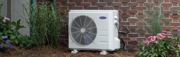 Outdoor air conditioner condenser unit against a brick wall, surrounded by mulch and flowering plants.