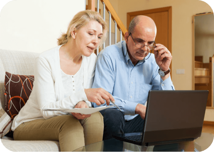 Older couple on a sofa reviewing a printed document while looking at a laptop at home.